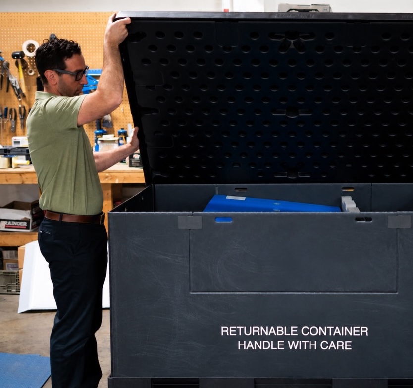 Worker opening a large black returnable container in an industrial workshop, with “Returnable Container Handle With Care” printed on the front